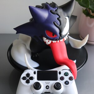 May include: A hybrid figure combining a purple and black creature with sharp teeth, a long red tongue, and a white eye, positioned behind a white video game controller featuring black analogue sticks, a directional pad, and four coloured action buttons.