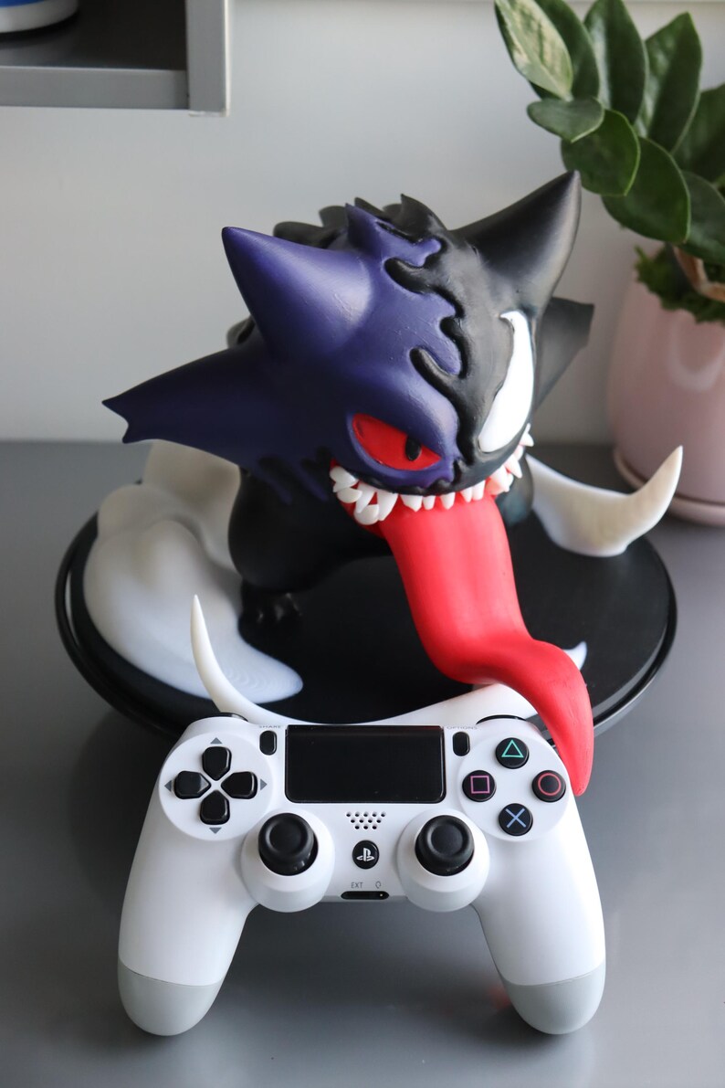 May include: A hybrid figure combining a purple and black creature with sharp teeth, a long red tongue, and a white eye, positioned behind a white video game controller featuring black analogue sticks, a directional pad, and four coloured action buttons.