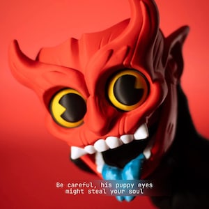May include: A red devil-like figurine with large yellow eyes, black pupils, white teeth, and a blue tongue. The text "Be careful, his puppy eyes might steal your soul" is printed at the bottom. The figure features prominent horns and ears.