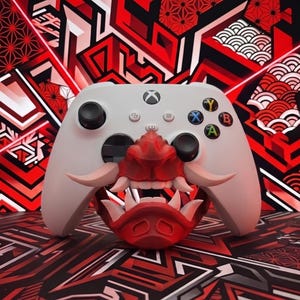 Könnte beinhalten: Ein weißer kabelloser Videospiel-Controller mit einem rot-weißen Maskenaufsatz im Samurai-Stil, der den mittleren Bereich bedeckt. Der Controller verfügt über schwarze Analogsticks, ein Steuerkreuz und farbige Aktionstasten.