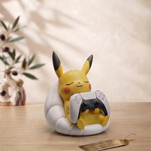 Puede incluir: Una figura amarilla de Pikachu con orejas de puntas negras y mejillas rojas, sentada en una silla blanca con forma de nube mientras sostiene un mando de videojuego blanco y negro. Una pequeña etiqueta metálica dorada con texto yace delante.