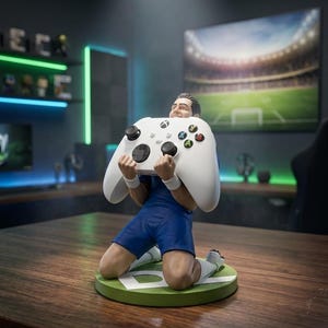 以下が含まれることがあります： 青いユニフォームを着たサッカー選手のフィギュアが緑色の円形ベースの上に跪き、大きな白いゲームコントローラーを抱えています。選手は白いリストバンドと白いソックスを着用しています。ベースには白い文字と「THE FIELD COMMANDER」というテキストが記されています。