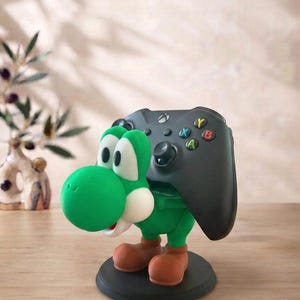 May include: A green Yoshi character figurine serving as a controller stand, featuring a black gaming controller resting on its back. The figure has a round white snout, large white eyes, and brown shoes, all mounted on a circular black base.