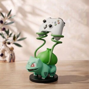 Puede incluir: Una figura de Bulbasaur de color verde claro sobre una base circular negra, con dos enredaderas verdes en espiral que se extienden hacia arriba para sostener un mando de videojuego inalámbrico blanco con joysticks negros y botones de colores.