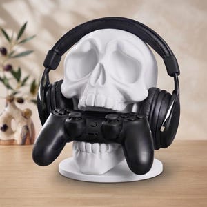 May include: A matte white skull-shaped stand designed to hold gaming accessories. The skull is wearing black over-ear headphones and has a black video game controller positioned horizontally across its jaw area, resting on a circular base.