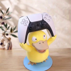 May include: A yellow duck-like character figurine standing on a light blue circular base, holding a white and black wireless game controller above its head with both arms raised. The character has a wide beak, small eyes, and a rounded body.