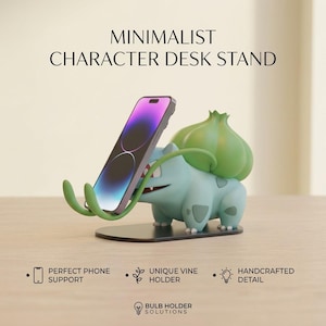 May include: A light blue and green creature-shaped desk stand holding a smartphone. The stand features a bulbous plant-like structure on its back and two curved vine-like arms to support the device. Text reads MINIMALIST CHARACTER DESK STAND.