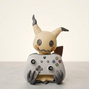 Puede incluir: Una figura beige de Mimikyu con puntas de orejas gris oscuro y detalles de ojos cosidos que sostiene un mando inalámbrico Xbox gris claro. El mando cuenta con una cruceta negra, dos joysticks negros, un botón central con el logo de Xbox y cuatro botones de acción de colores etiquetados como Y, B, A y X.