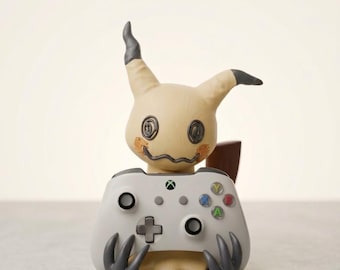 May include: A beige Mimikyu figure with dark grey ear tips and stitched eye details holding a light grey Xbox wireless controller. The controller features a black directional pad, two black analogue sticks, a central Xbox logo button, and four coloured action buttons labelled Y, B, A, and X.