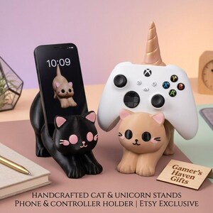Op de afbeelding: Twee katvormige standaards, een zwarte en een beige, ontworpen om een smartphone en een witte gamecontroller vast te houden. De beige kat heeft een gouden eenhoornhoorn. Een klein houten bordje leest Gamer's Haven Gifts. De tekst onderaan leest HANDCRAFTED CAT & UNICORN STANDS PHONE & CONTROLLER HOLDER | ETSY EXCLUSIVE.