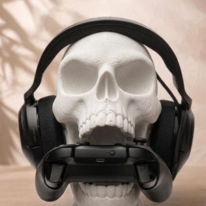 May include: A white 3D-printed human skull model wearing black over-ear headphones. The skull has an open jaw holding a black video game controller between its teeth. The surface of the skull shows visible horizontal layer lines from the printing process.