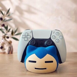 May include: A white and black video game controller resting on a blue and cream-colored Snorlax-shaped plastic stand. The stand features the character's signature closed eyes, small ears, and a wide, flat face design, providing a stable base for the controller.