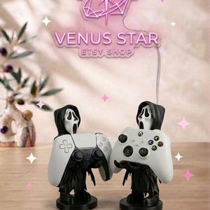 May include: Two black and white ghost-themed controller stands, each featuring a hooded figure with a white mask, holding a white gaming controller. Above them, a pink neon sign displays the text VENUS STAR ETSY SHOP with a star icon.