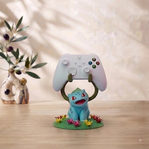 May include: A light blue Bulbasaur-shaped controller stand holding a white Xbox wireless controller. The base is green with small yellow and pink flowers and features the text "HANDCRAFTED BY TRAINER CRAFTS" printed along the front edge.