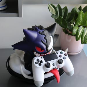 May include: A purple and black creature figurine with sharp teeth and red eyes, featuring a split design, holding a white video game controller with a long red tongue extending from its mouth, all mounted on a circular black base.
