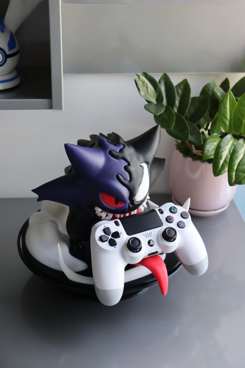 May include: A purple and black creature figurine with sharp teeth and red eyes, featuring a split design, holding a white video game controller with a long red tongue extending from its mouth, all mounted on a circular black base.