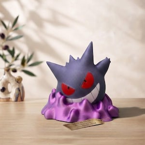 Può includere: Una statuetta di Gengar viola con orecchie a punta e punte, caratterizzata da occhi rossi luminosi e un ampio sorriso bianco. Il personaggio emerge da una base viola lucida e strutturata che imita un effetto liquido, con una piccola etichetta beige.