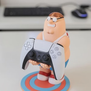 May include: A figurine of a cartoon character with brown hair and glasses, wearing a white shirt and blue shorts, standing on a circular orange and blue base. The character is holding a white and black video game controller in its hands.