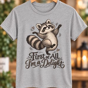 Puede incluir: Una camiseta gris jaspeado de manga corta con una ilustración gráfica de un mapache bailando con una cola a rayas. Debajo del mapache, el texto First of All I'm a Delight aparece en una fuente cursiva oscura y estilizada con corazones.