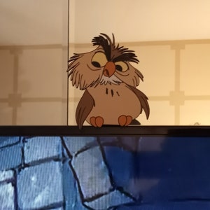 May include: A brown and white cartoon owl with a grumpy expression is perched on top of a television screen. The television screen is showing a scene of a stone wall.