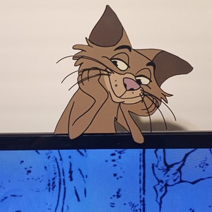 May include: A flat, brown, cartoon-style cat cutout with dark brown ear tips and a pink nose. The cat is depicted with its head resting on one paw, featuring long black whiskers and a smug expression, designed to perch on the top edge of a monitor.