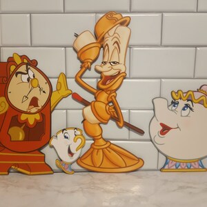 May include: Four characters from the Disney film Beauty and the Beast. The characters are a red clock, a yellow candlestick, a white teapot, and a white teacup. The characters are all smiling and looking at the viewer.