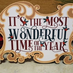 Puede incluir: Un letrero decorativo con el texto "IT'S THE MOST WONDERFUL TIME OF THE YEAR" en borgo&ntilde;a y verde oscuro. El letrero tiene un marco dorado decorativo y acentos de acebo. Mickey Mouse es visible a la izquierda.