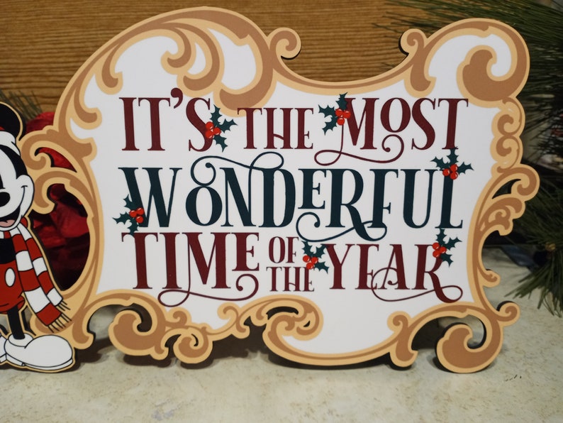 Puede incluir: Un letrero decorativo con el texto "IT'S THE MOST WONDERFUL TIME OF THE YEAR" en borgo&ntilde;a y verde oscuro. El letrero tiene un marco dorado decorativo y acentos de acebo. Mickey Mouse es visible a la izquierda.