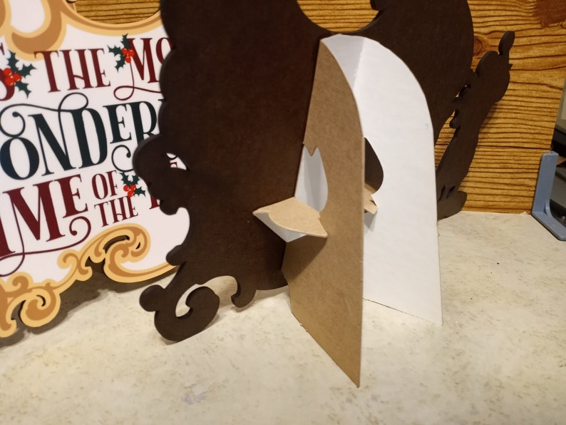 Puede incluir: Un letrero decorativo marr&oacute;n con texto blanco y rojo que dice "THE MOST WONDERFUL TIME OF THE YEAR". El letrero tiene un soporte de cart&oacute;n adjunto en la parte posterior.