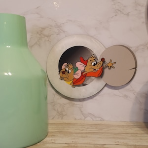 May include: A white and gray round wall decor with a hole in the center. Two cartoon mice are peeking out of the hole. The mouse on the left is brown and the mouse on the right is brown with a red hat and a red shirt.
