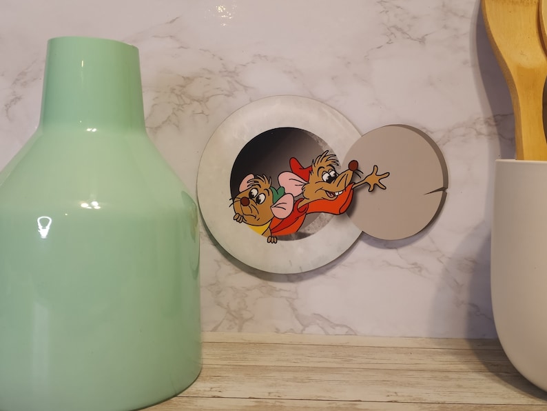 May include: A white and gray round wall decor with a hole in the center. Two cartoon mice are peeking out of the hole. The mouse on the left is brown and the mouse on the right is brown with a red hat and a red shirt.