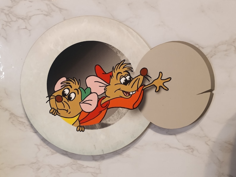 May include: Two cartoon mice peeking out of a hole in a white circle. The mouse on the left is brown and wearing a green hat. The mouse on the right is brown and wearing a red and orange hat. The background is a white marble surface.