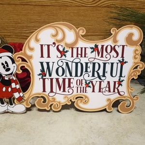 Puede incluir: Un letrero de madera con tem&aacute;tica navide&ntilde;a con la frase "IT'S THE MOST WONDERFUL TIME OF THE YEAR" en rojo, blanco y verde oscuro. El letrero est&aacute; decorado con acebo y una imagen de dibujos animados de Mickey Mouse con gorro y bufanda de Pap&aacute; Noel.