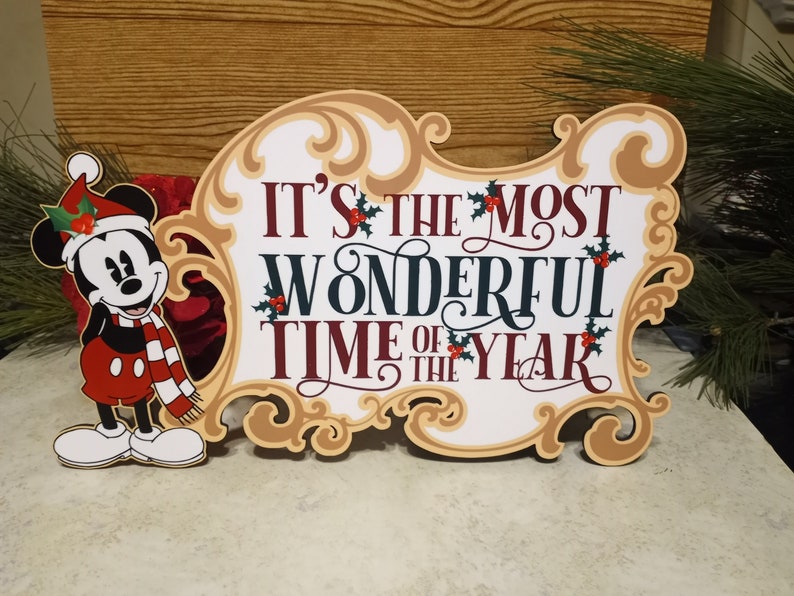 Puede incluir: Un letrero de madera con tem&aacute;tica navide&ntilde;a con la frase "IT'S THE MOST WONDERFUL TIME OF THE YEAR" en rojo, blanco y verde oscuro. El letrero est&aacute; decorado con acebo y una imagen de dibujos animados de Mickey Mouse con gorro y bufanda de Pap&aacute; Noel.