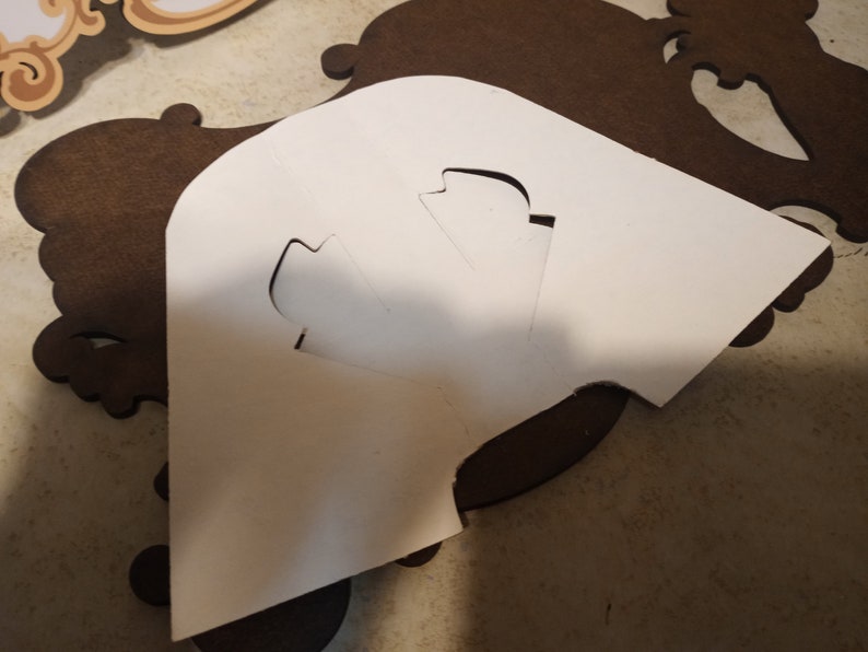 Puede incluir: Una plantilla de papel blanco con formas recortadas se coloca sobre una pieza decorativa de madera marr&oacute;n oscuro. La pieza de madera tiene un dise&ntilde;o ornamental y enrollado. La plantilla parece ser para un proyecto de manualidades.