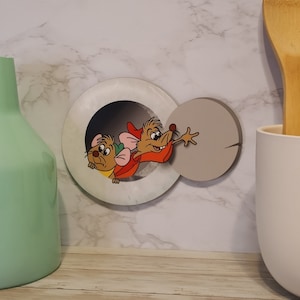 May include: A white and gray wooden wall decor featuring two cartoon mice peeking out of a round hole. The mice are brown and have red accents.