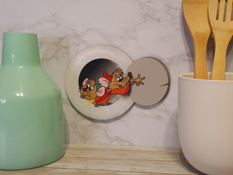 May include: A white and gray wooden wall decor featuring two cartoon mice peeking out of a round hole. The mice are brown and have red accents.