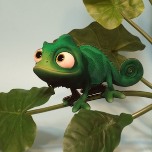 Puede incluir: Figura de camaleón verde con ojos grandes y expresivos, posada en una vid falsa con hojas verdes grandes. El camaleón tiene piel texturizada y una cola enroscada. El fondo es azul claro.