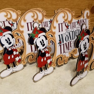 Puede incluir: Varias decoraciones navide&ntilde;as de Mickey Mouse. Cada una presenta a Mickey con gorro y bufanda de Pap&aacute; Noel, frente a un letrero que dice "IT'S THE MOST WONDERFUL TIME OF THE YEAR" en texto rojo. Los letreros tienen un borde dorado ornamentado.