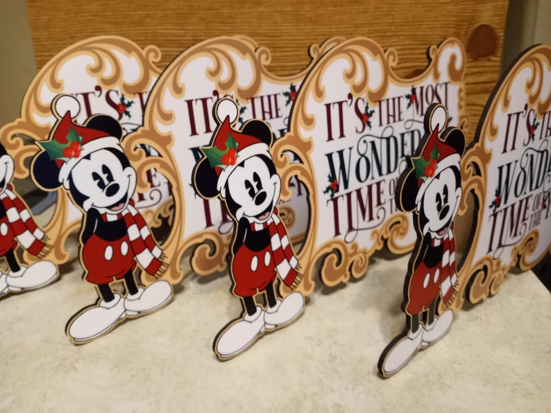 Puede incluir: Varias decoraciones navide&ntilde;as de Mickey Mouse. Cada una presenta a Mickey con gorro y bufanda de Pap&aacute; Noel, frente a un letrero que dice "IT'S THE MOST WONDERFUL TIME OF THE YEAR" en texto rojo. Los letreros tienen un borde dorado ornamentado.