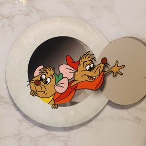 May include: Two cartoon mice peeking out of a round hole. The mouse on the left is brown with a green hat and pink ears. The mouse on the right is brown with a red hat and is reaching out with its right paw.