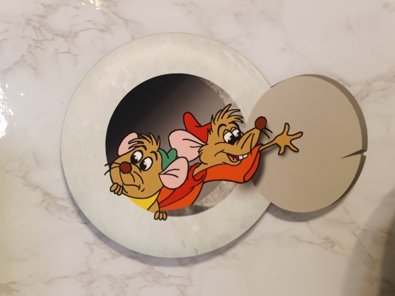 May include: Two cartoon mice peeking out of a round hole. The mouse on the left is brown with a green hat and pink ears. The mouse on the right is brown with a red hat and is reaching out with its right paw.