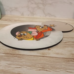May include: A round, white coaster with a black border features a cartoon image of two mice peeking out of a hole. The mouse on the left is brown with a green hat and the mouse on the right is brown with a red shirt.