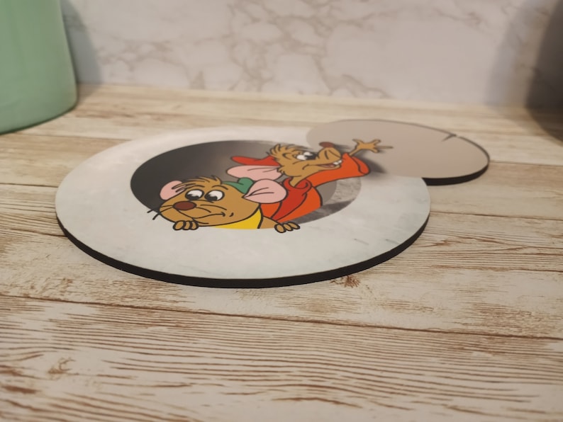 May include: A round, white coaster with a black border features a cartoon image of two mice peeking out of a hole. The mouse on the left is brown with a green hat and the mouse on the right is brown with a red shirt.