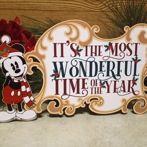 Puede incluir: Un letrero navide&ntilde;o festivo con Mickey Mouse con gorro y bufanda de Pap&aacute; Noel. El letrero dice "It's the Most Wonderful Time of the Year" en una fuente elegante, con detalles de acebo. El letrero tiene un marco dorado.