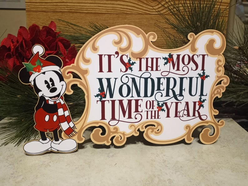 Puede incluir: Un letrero navide&ntilde;o festivo con Mickey Mouse con gorro y bufanda de Pap&aacute; Noel. El letrero dice "It's the Most Wonderful Time of the Year" en una fuente elegante, con detalles de acebo. El letrero tiene un marco dorado.