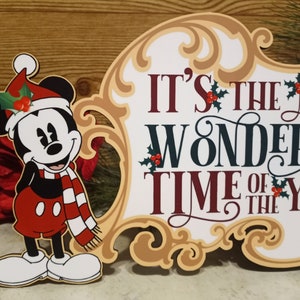 Puede incluir: Un letrero navide&ntilde;o con Mickey Mouse con gorro y bufanda de Pap&aacute; Noel. El letrero dice "It's the Most Wonderful Time of the Year" en colores festivos, con acentos de acebo. El letrero tiene un borde ornamentado.