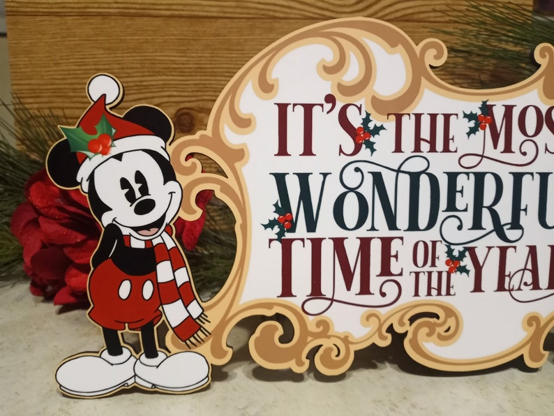 Puede incluir: Un letrero navide&ntilde;o con Mickey Mouse con gorro y bufanda de Pap&aacute; Noel. El letrero dice "It's the Most Wonderful Time of the Year" en colores festivos, con acentos de acebo. El letrero tiene un borde ornamentado.
