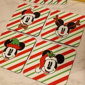 Può includere: Set di sottobicchieri quadrati con Topolino in abiti natalizi. Ogni sottobicchiere ha uno sfondo a righe rosse e verdi. Mickey indossa un cappello da Babbo Natale, un cappello da elfo, corna di renna e un cappello da Babbo Natale con agrifoglio.