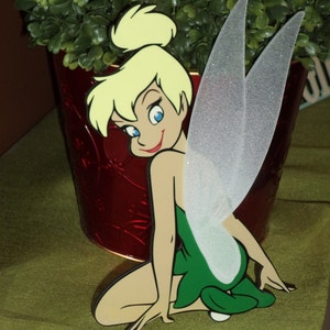 Puede incluir: Una figurita de Tinkerbell verde y blanca con cabello rubio y un vestido verde. Está sentada con las alas extendidas detrás de ella.